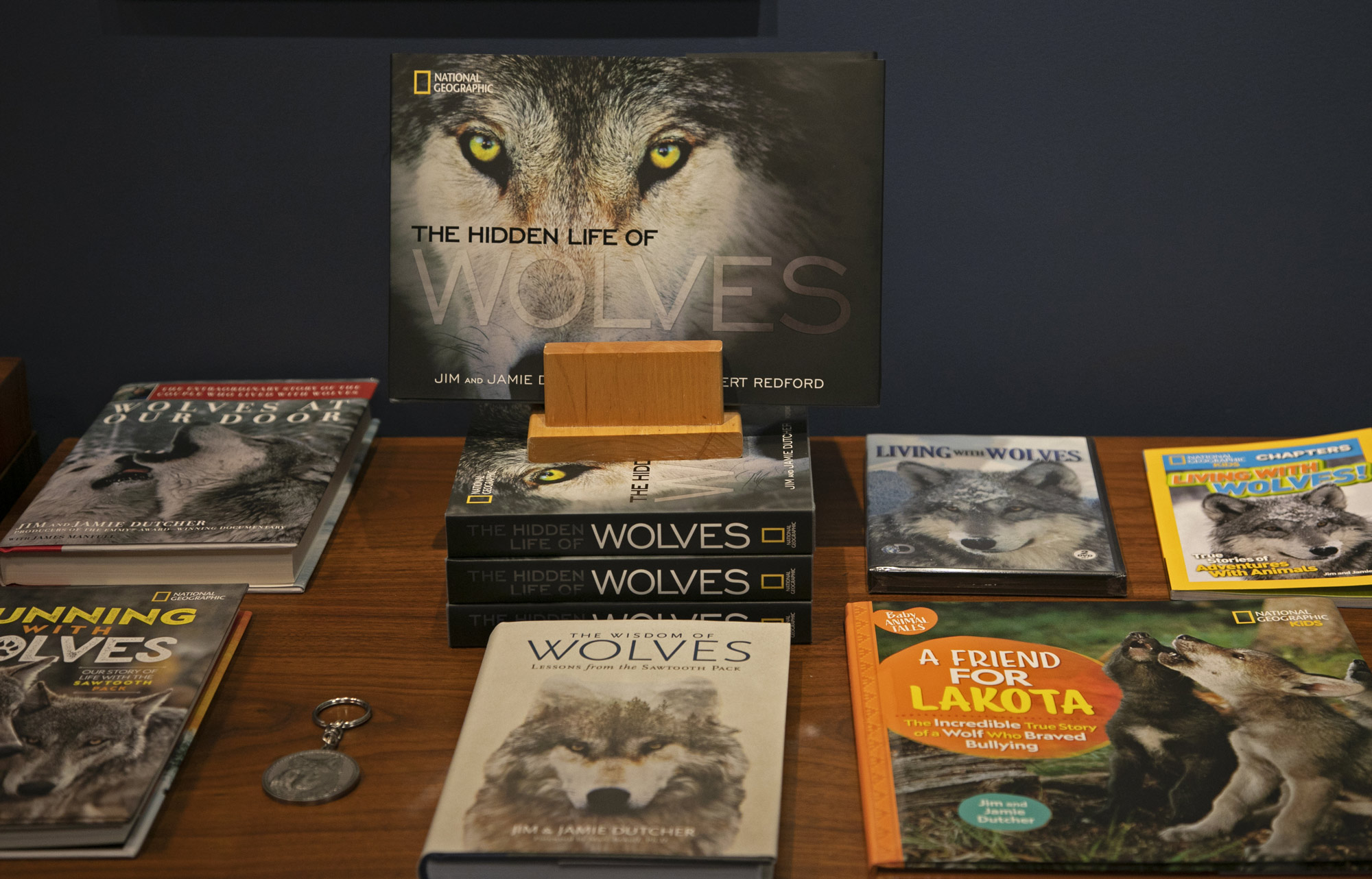 A display of wolf-themed books and magazines, including “The Hidden Life of Wolves” by National Geographic, arranged on a wooden table with a keychain—showcasing what we do to celebrate these remarkable creatures. | Living with Wolves