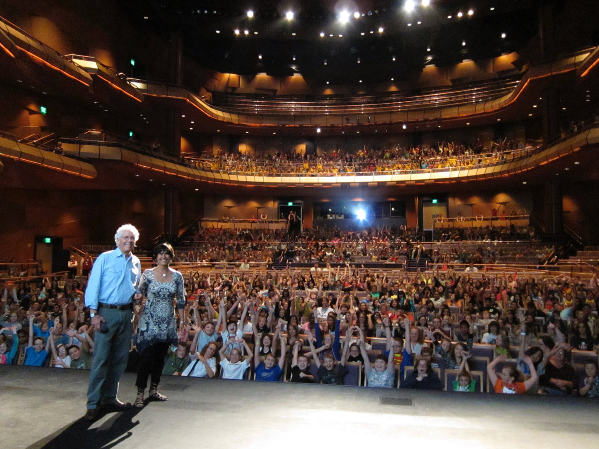 A man and a woman stand on stage facing a packed auditorium filled with people. Showcasing what we do, the audience on multiple levels raises their hands enthusiastically toward the stage. | Living with Wolves