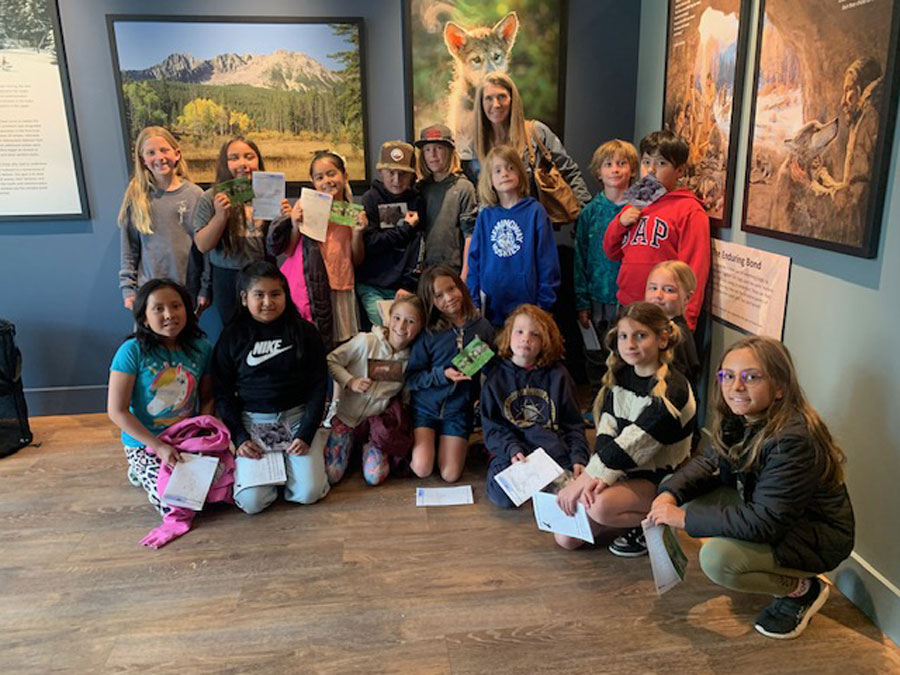 A group of children and an adult pose inside the Living with Wolves museum, some holding papers or booklets. There is a large wolf photo on the wall behind them. Everyone is smiling at the camera. | Living with Wolves