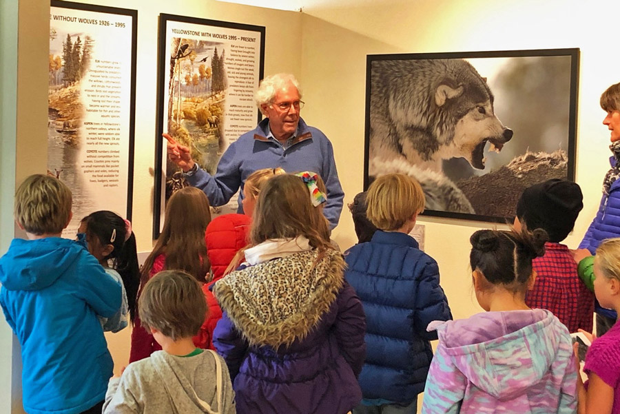 An older man explains "What we do" at a museum exhibit about wolves to children in colorful jackets, as large wolf photos and informational posters fill the wall behind him. | Living with Wolves