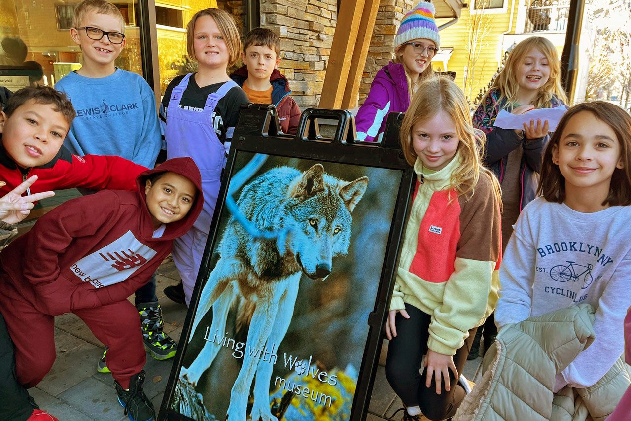 A group of smiling children stand around a display board featuring an image of a wolf and the text Living with Wolves Museum, showing what we do to educate and inspire. The kids are wearing colorful casual clothes and jackets, posing outdoors. | Living with Wolves