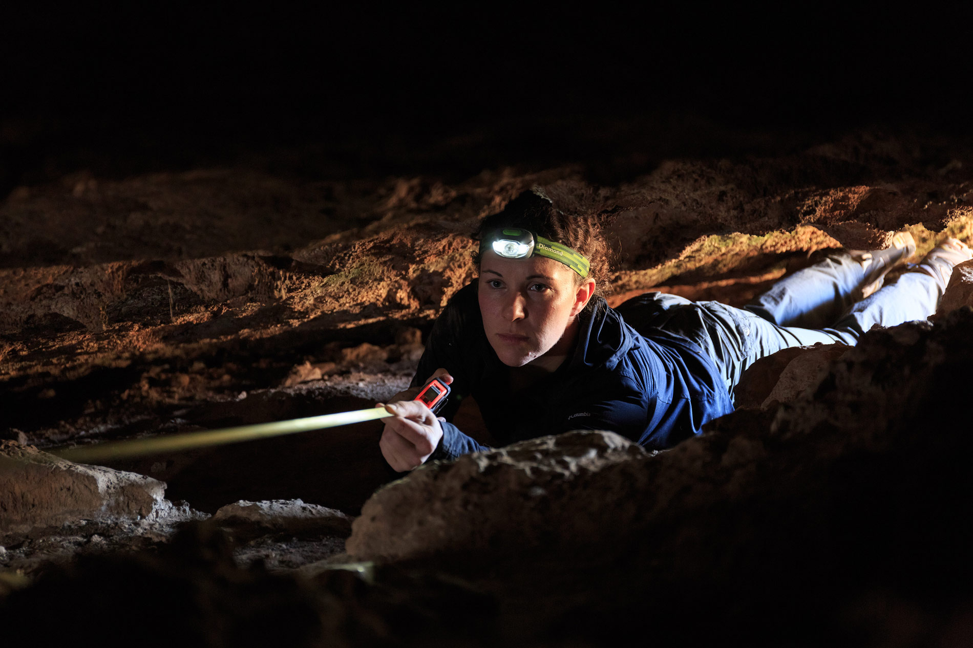 A person wearing a headlamp crawls through a narrow cave passage, using a measuring tape to record dimensions—showcasing what we do as we carefully explore and document confined spaces amid rocks and dim lighting. | Living with Wolves
