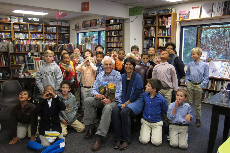 A group of children poses with two adults seated in a bookstore, with bookshelves in the background. Some smile while others make silly faces, showcasing what we do to create a lively and joyful atmosphere. | Living with Wolves